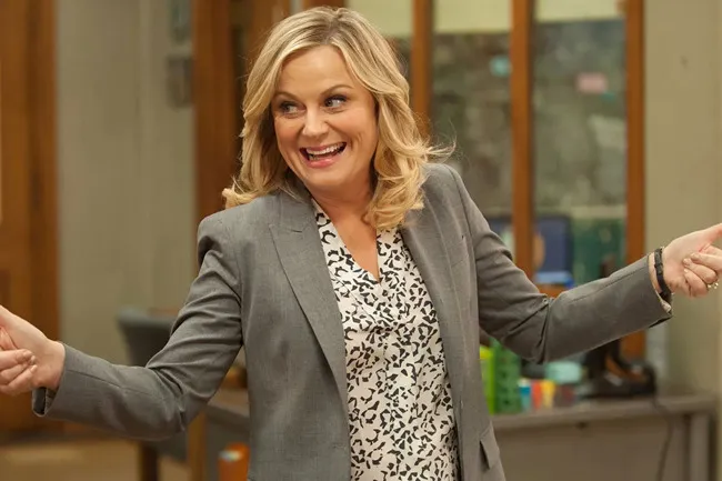 Actress in a gray blazer and patterned blouse smiling happily in a scene from Parks and Recreation comfort TV rerun.