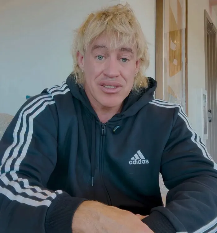 Influencer with blonde hair wearing black Adidas hoodie sitting indoors in a casual setting, appearing cringy and unapologetic.