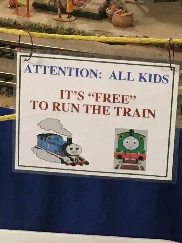 Sign with suspicious quotation marks saying kids can "free" run the train, featuring cartoon train images at a model train display.