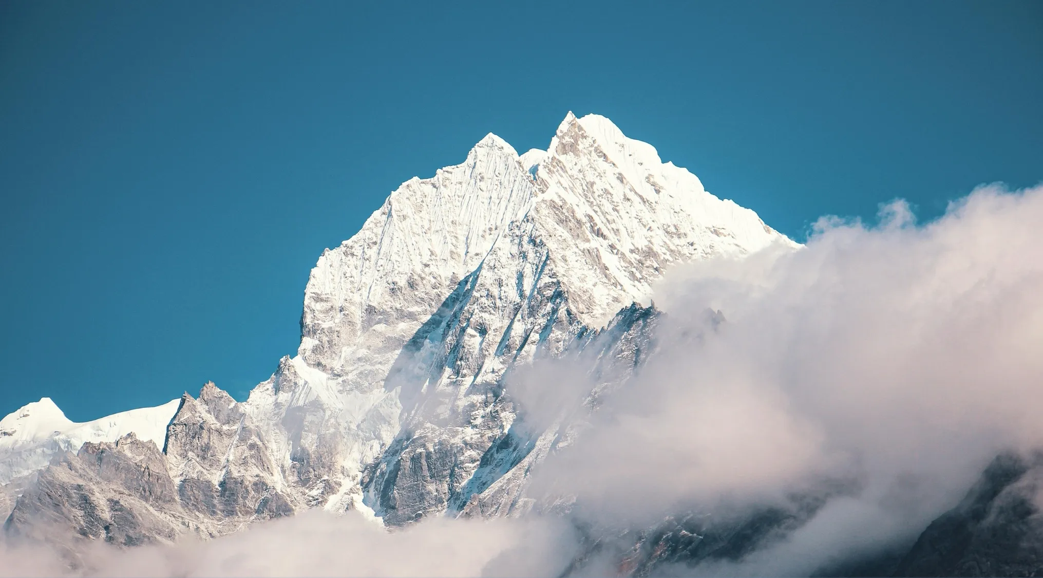 Snow-covered mountain peak rising above clouds under a clear blue sky in a geography edition setting.