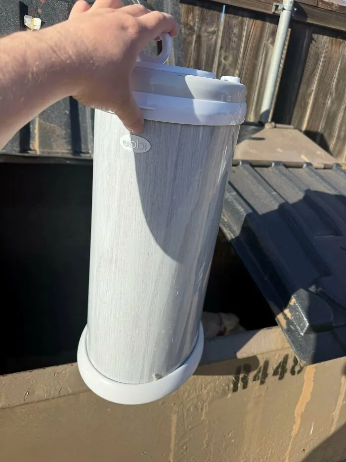 A hand holds a baby diaper pail over a dumpster, representing the funny side of being a great dad.