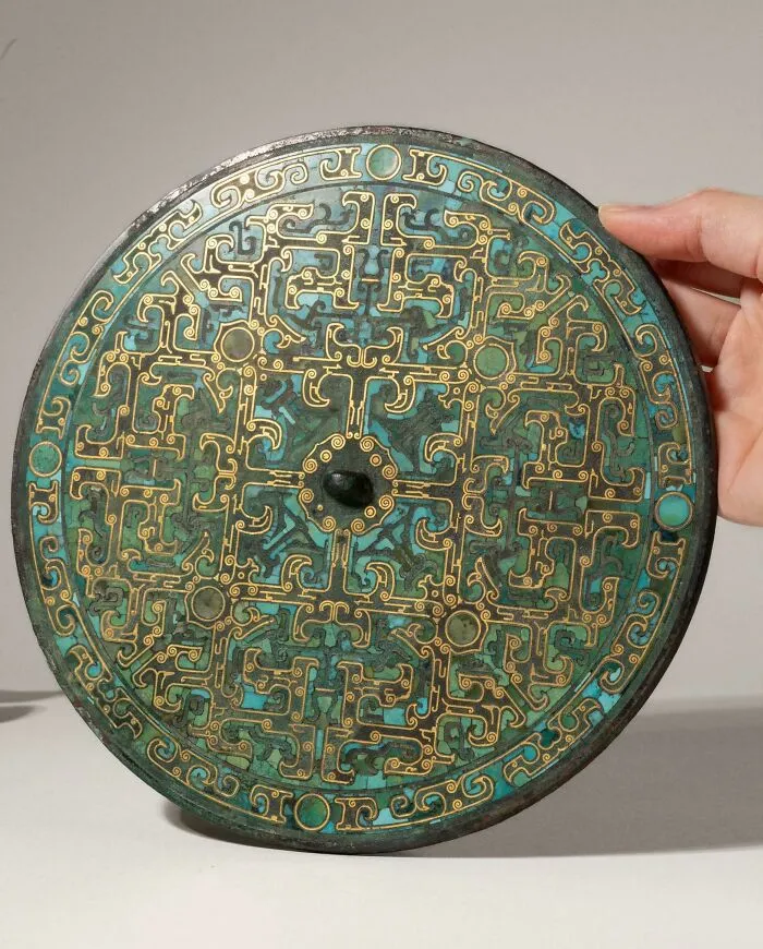 A hand holding an intricately detailed ancient bronze mirror with turquoise inlay from historical finds.
