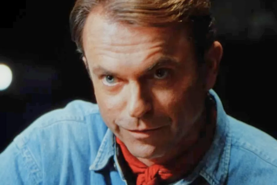 Jurassic Park character in a blue shirt and red scarf, looking intently with a slight smile in a dimly lit scene.