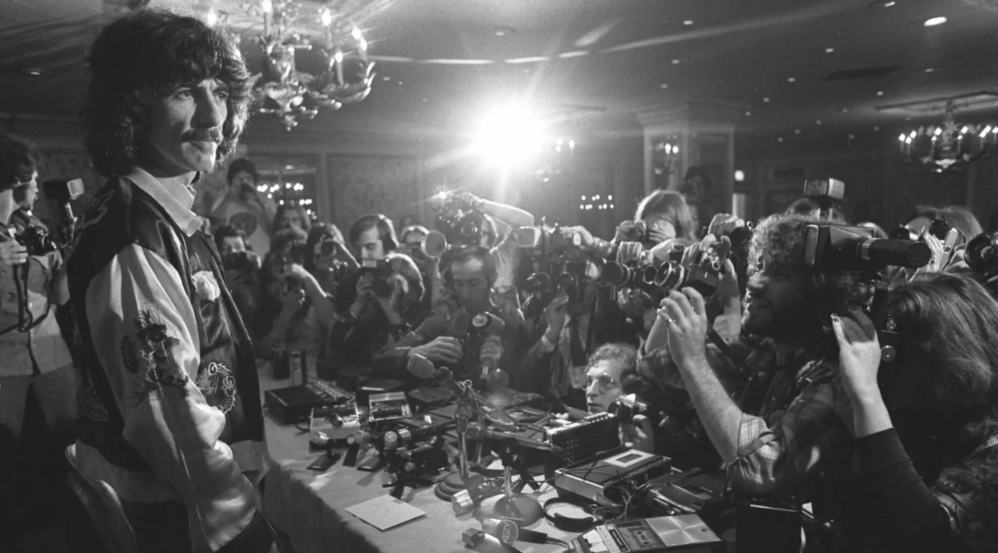 George Harrison at a press conference surrounded by photographers capturing images and conducting interviews.