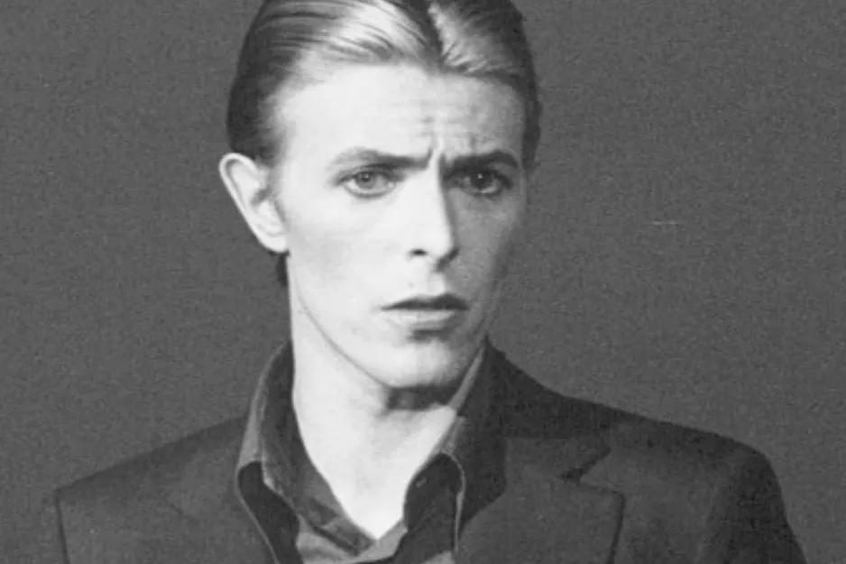 Black and white portrait of David Bowie wearing a suit and looking seriously into the camera in a studio setting.