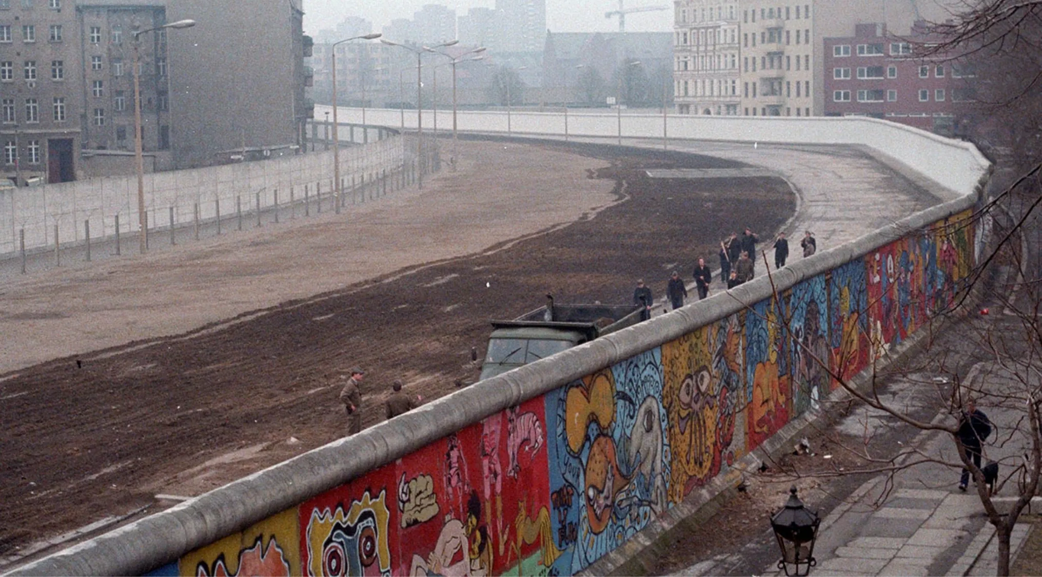 Section of the Berlin Wall covered in colorful graffiti with people walking nearby in the 1980s.