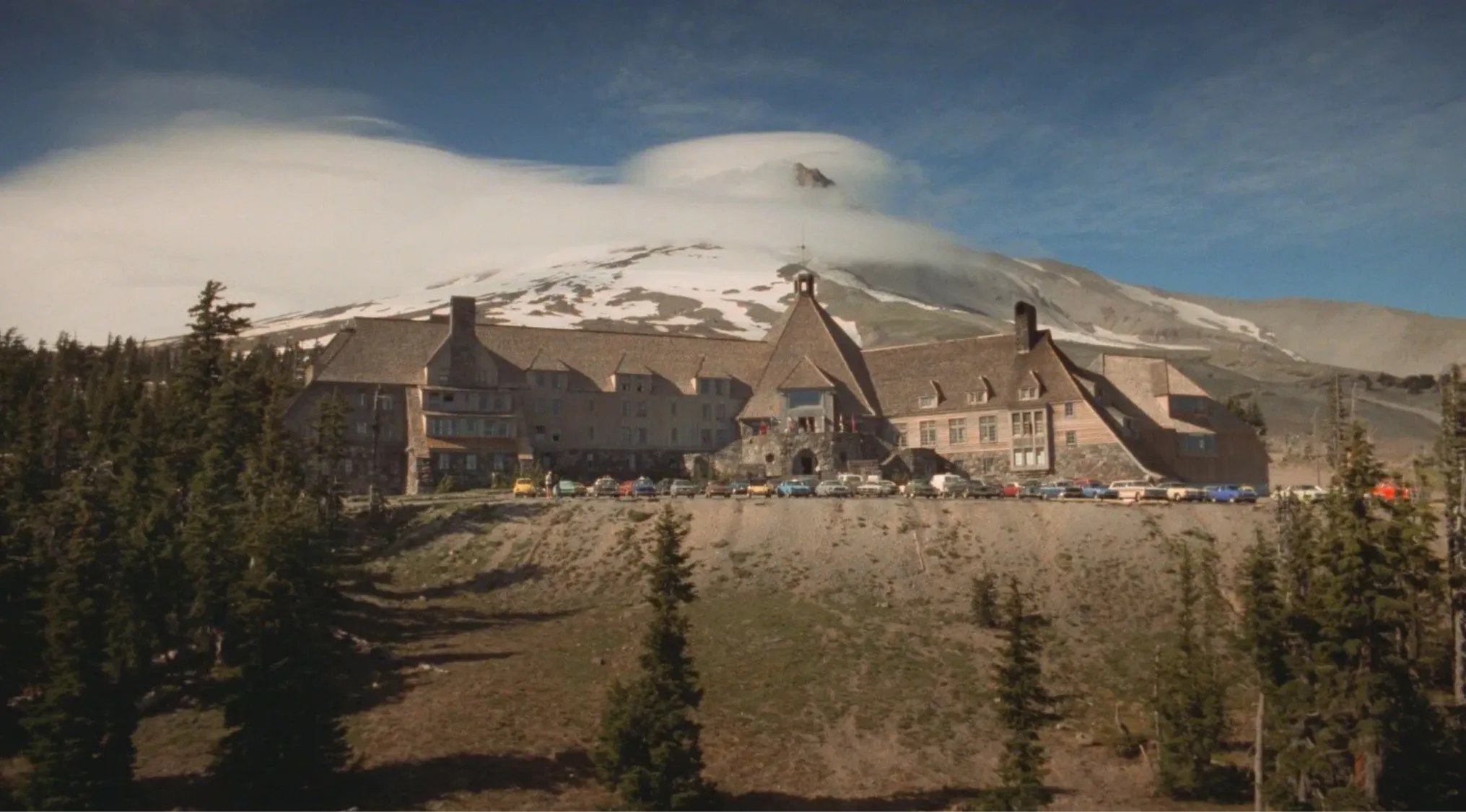 The Shining hotel exterior with mountain backdrop and classic cars parked, iconic 80s pop culture location.