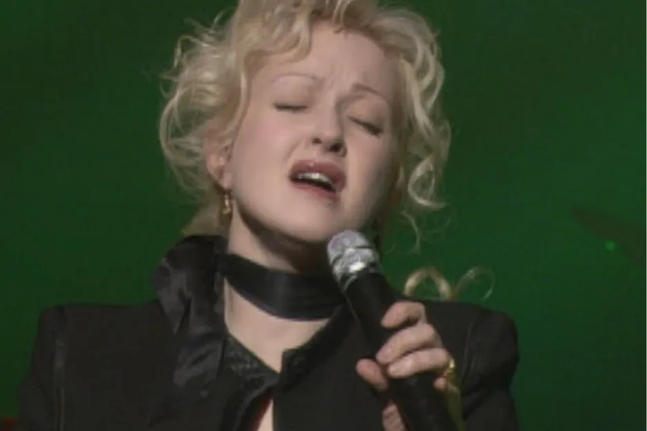 Cyndi Lauper performing on stage in the 80s, known as the Queen of Pop with blonde curly hair and black outfit.
