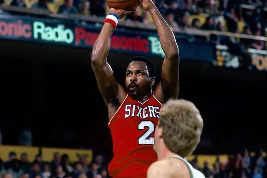 NBA player Moses Malone in Sixers uniform shooting basketball during a game with defender in foreground