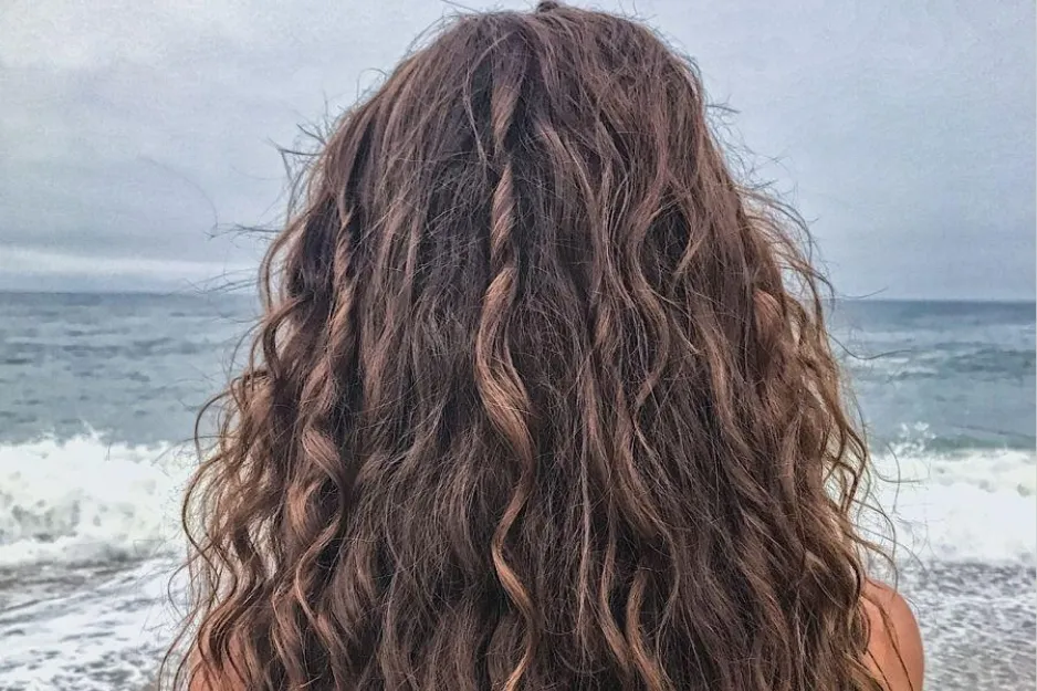Woman with natural beach waves hairstyle standing by the ocean, showcasing 1980s trend of wavy hair texture.