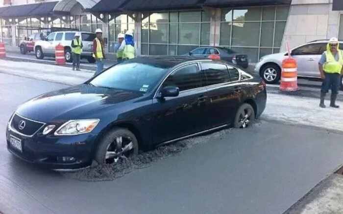 Black Lexus stuck in wet concrete on a street under construction, workers in safety vests standing nearby showing uncultured ignorance.