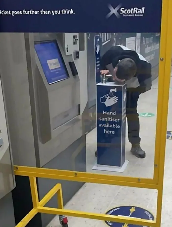 Man bending down to drink from a hand sanitizer dispenser in a station, highlighting uncultured and ignorant Americans.
