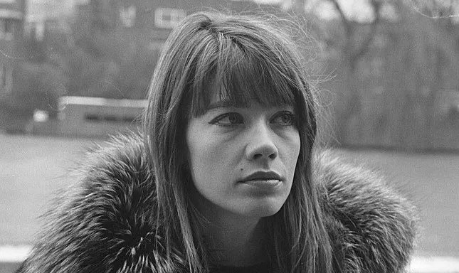 Black and white photo of Françoise Hardy looking contemplative outdoors with fur collar coat.