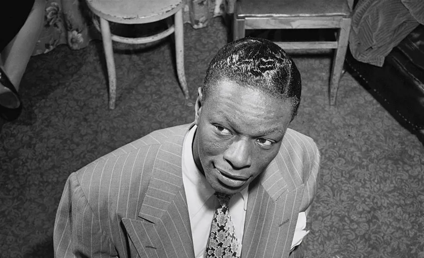 Nat King Cole in a pinstripe suit looking up, seated in a room with vintage chairs and patterned carpet.