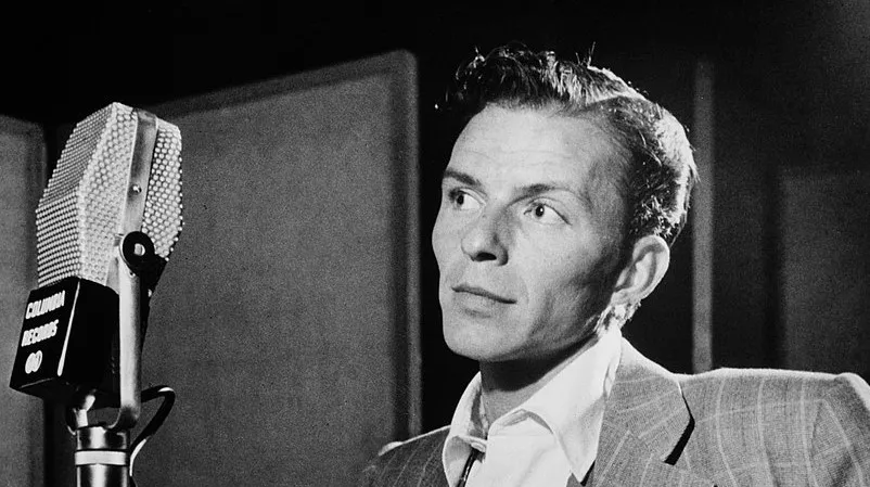 Frank Sinatra in a recording studio, wearing a suit, preparing to sing into a vintage Columbia Records microphone.
