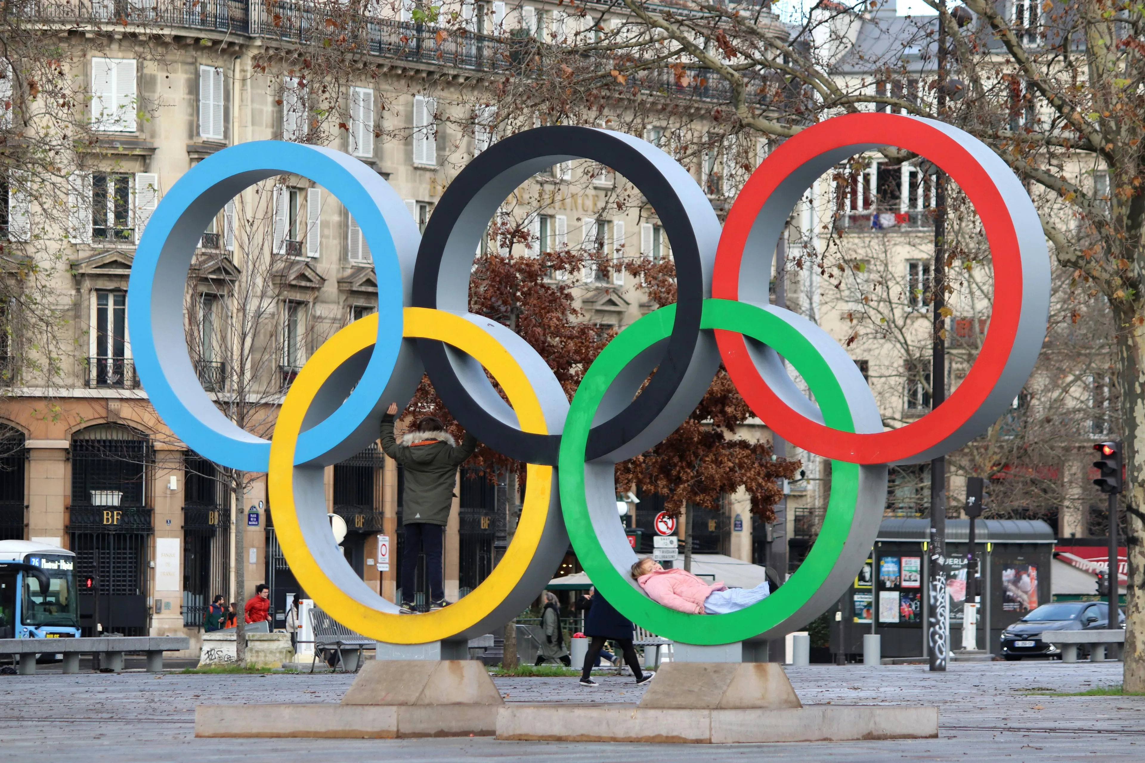 Large colorful Olympic rings sculpture in a city square with children playing inside the rings on a cloudy day.