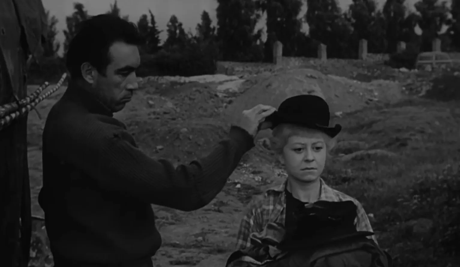Black and white scene from La Strada showing a man adjusting a hat on a woman, Italian film director and 1950s film setting.