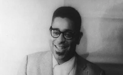 Black and white photo of a legendary jazz musician wearing glasses and a suit, smiling warmly in a classic portrait.