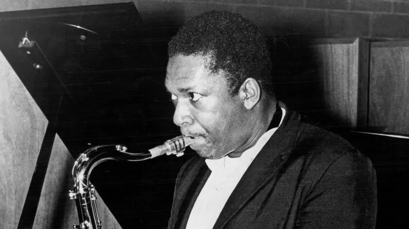 Legendary jazz musician John Coltrane playing saxophone in a black suit during a studio session.