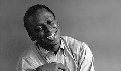 Miles Davis smiling in a casual shirt, black and white portrait highlighting the legendary jazz musician’s iconic presence.
