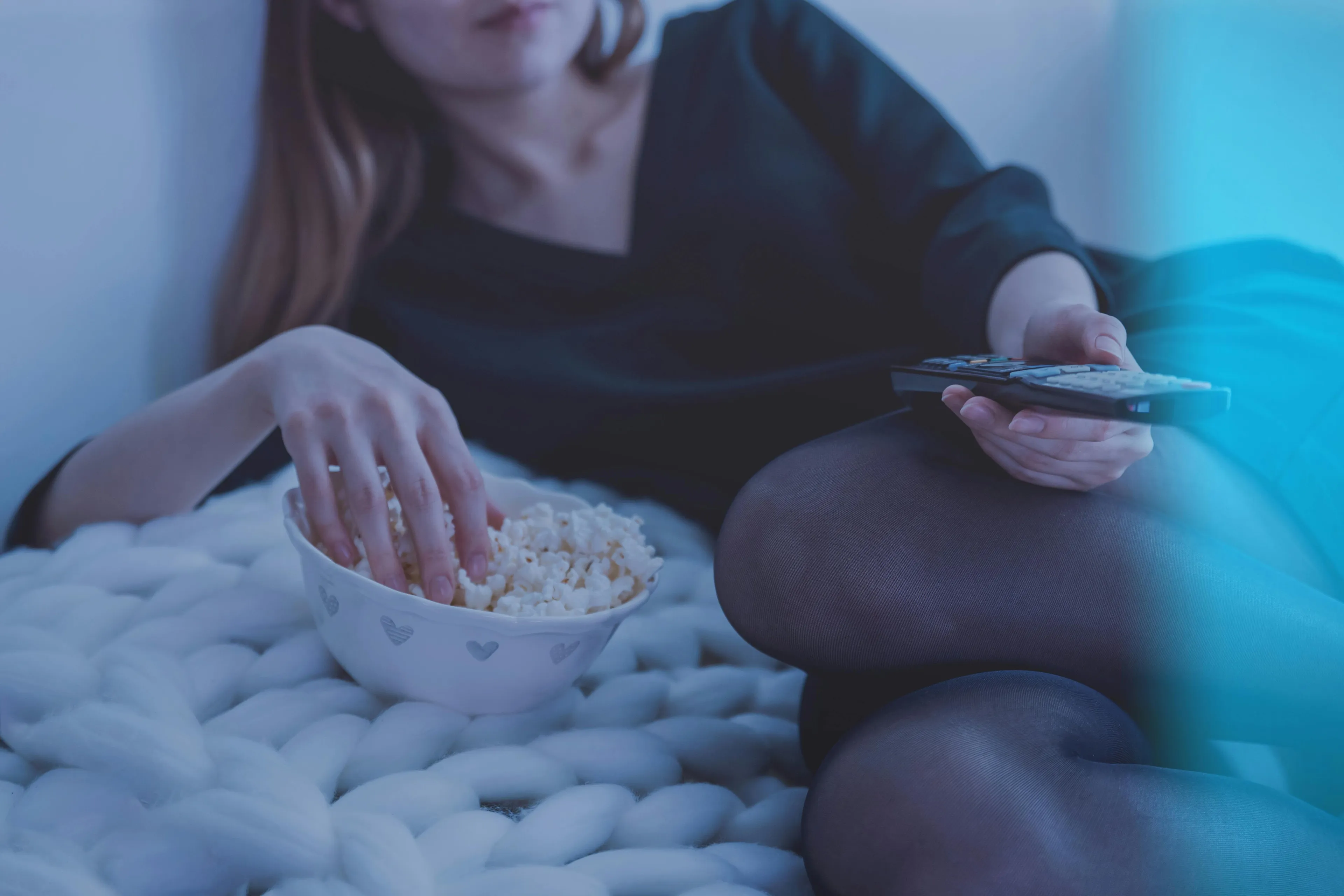 Person holding a remote and eating popcorn while relaxing on a cozy blanket during a movie night at home.