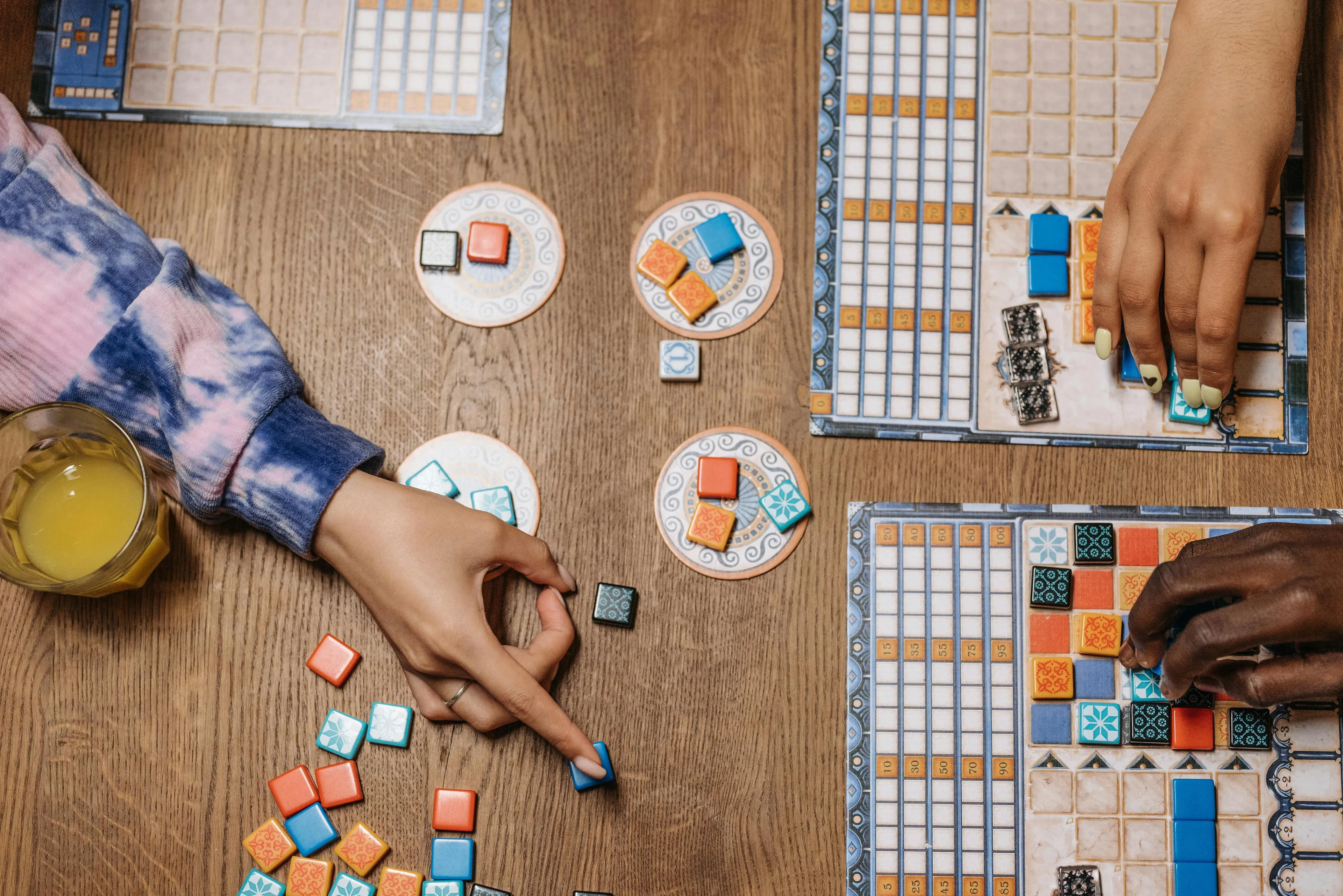 Hands of friends playing a board game night at a wooden table with colorful tiles and a glass of juice nearby