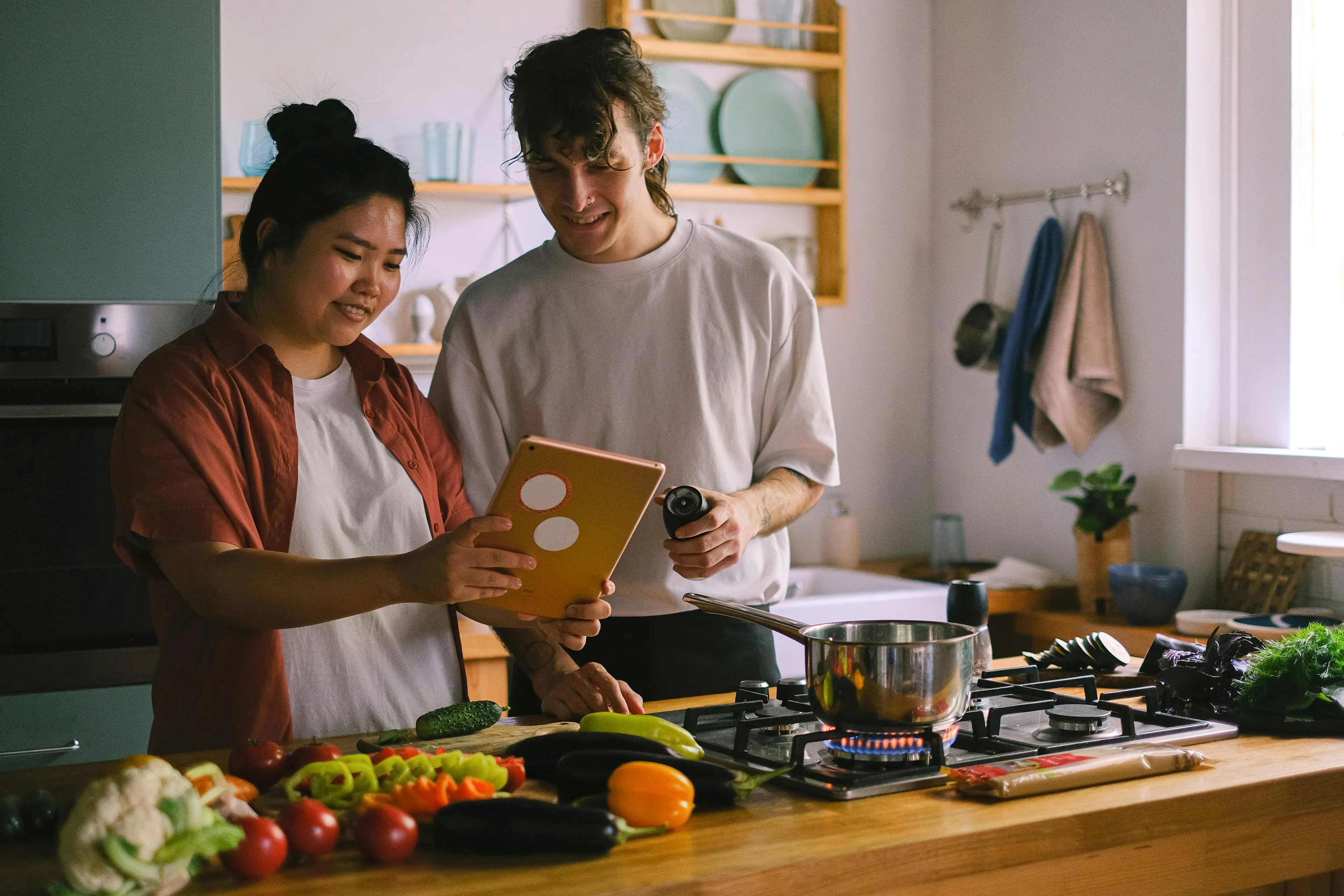 Two friends cooking dinner together in a kitchen, preparing fresh vegetables and using a tablet for recipes.