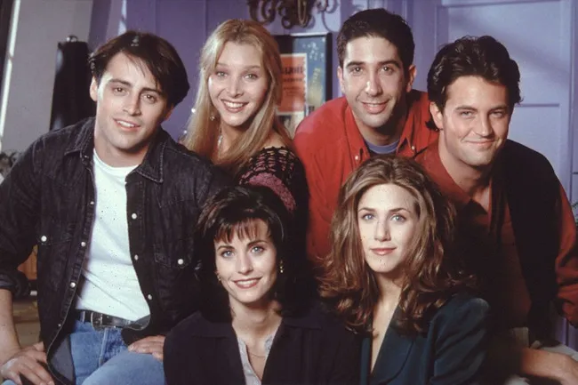 Cast of Friends sitcom posing together in a cozy apartment setting, showing iconic 90s fashion and hairstyles.