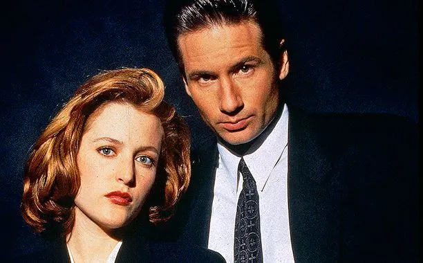 Actors from The X-Files standing closely together against a dark background, wearing professional suits and serious expressions.