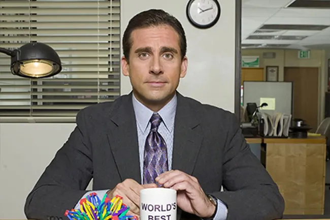 Michael Scott from The Office sitting at desk with World's Best Boss mug in an office setting.
