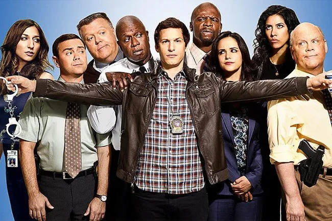 Cast of Brooklyn Nine-Nine posing together with detective in the center wearing a badge and leather jacket.