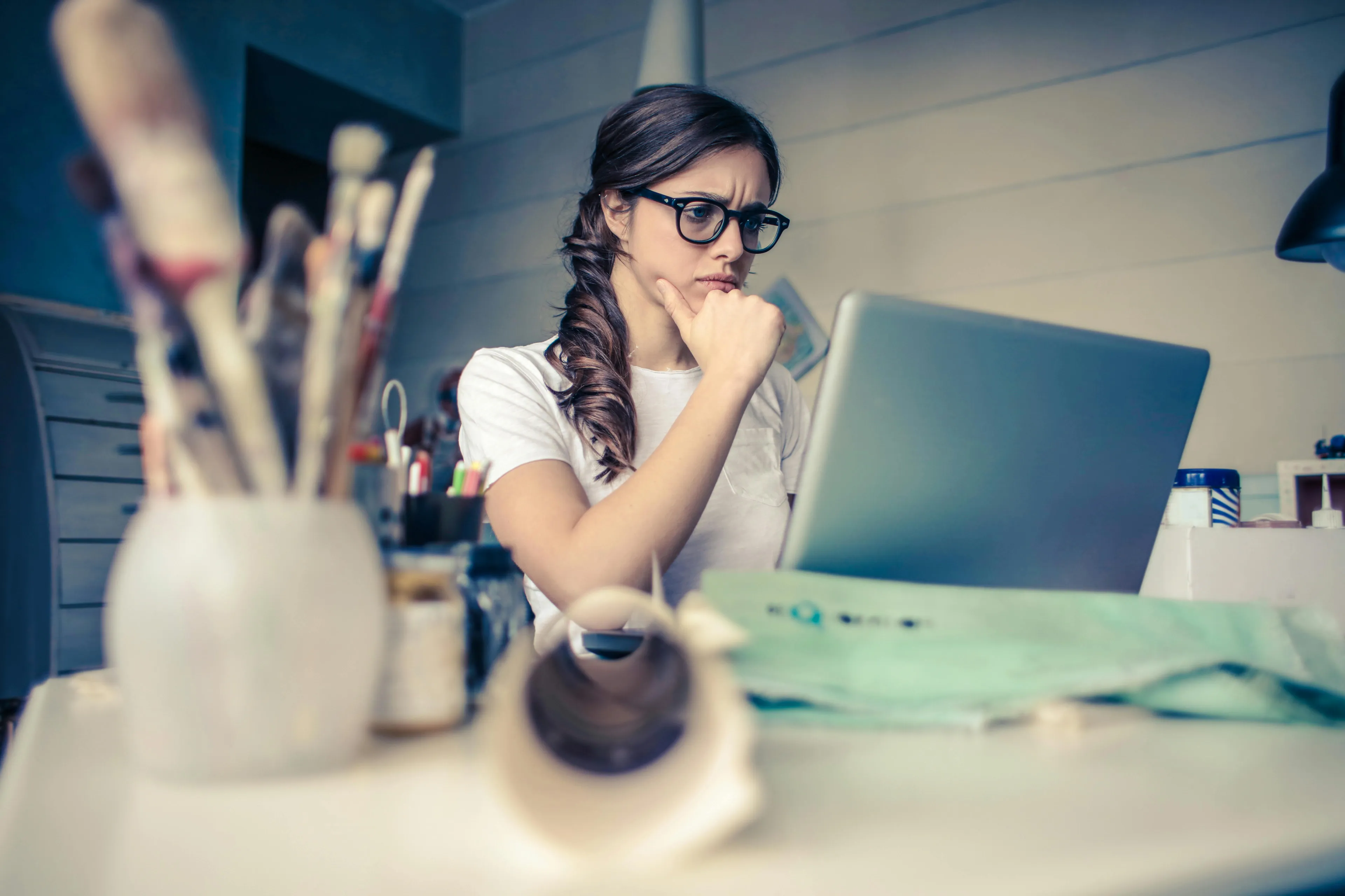 Young woman wearing glasses, trying to control urges while working on a laptop in a creative workspace.