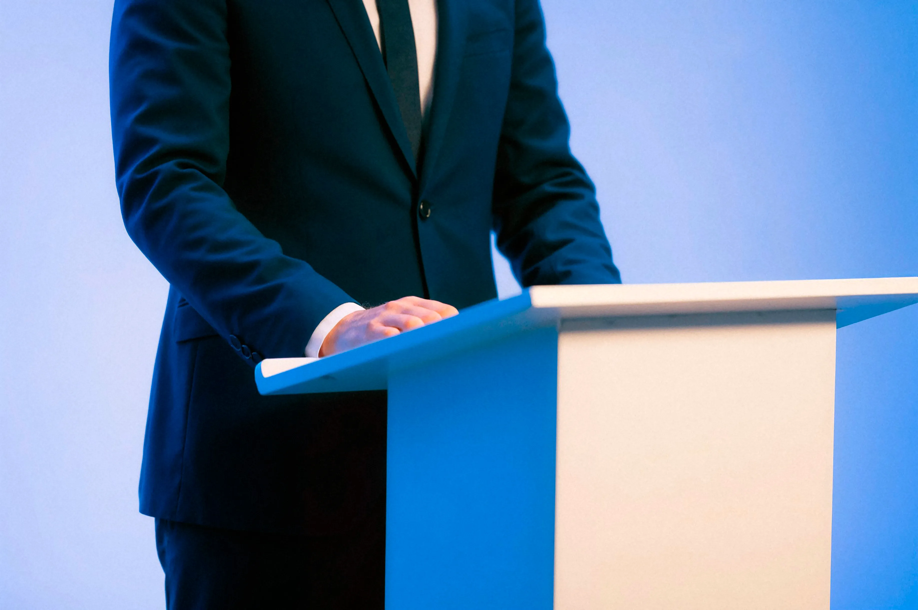 Man in a dark suit standing behind a white podium, participating in a political debate trending online discussion.