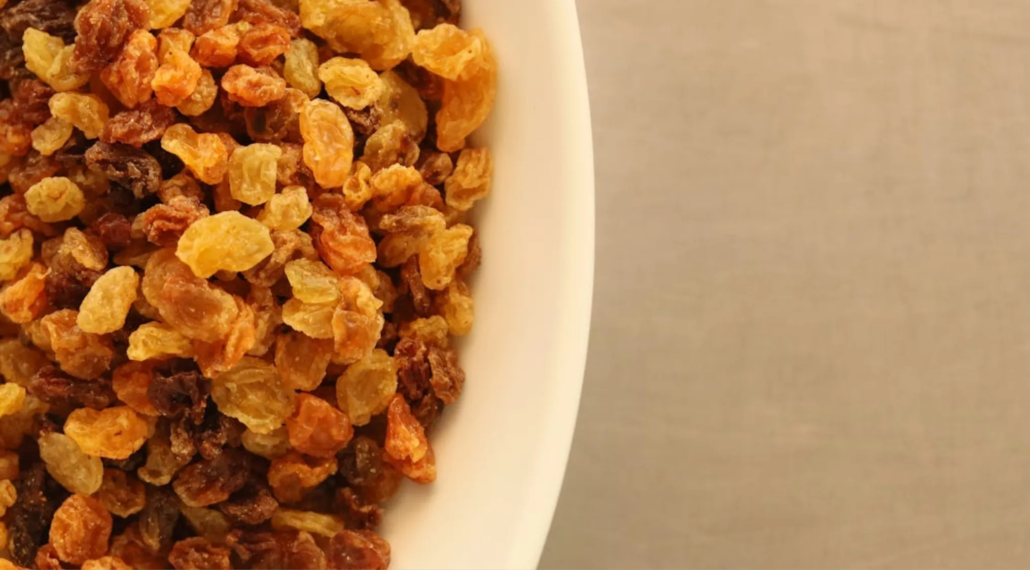 Bowl filled with golden and brown raisins made from dried grapes on a light brown surface background.