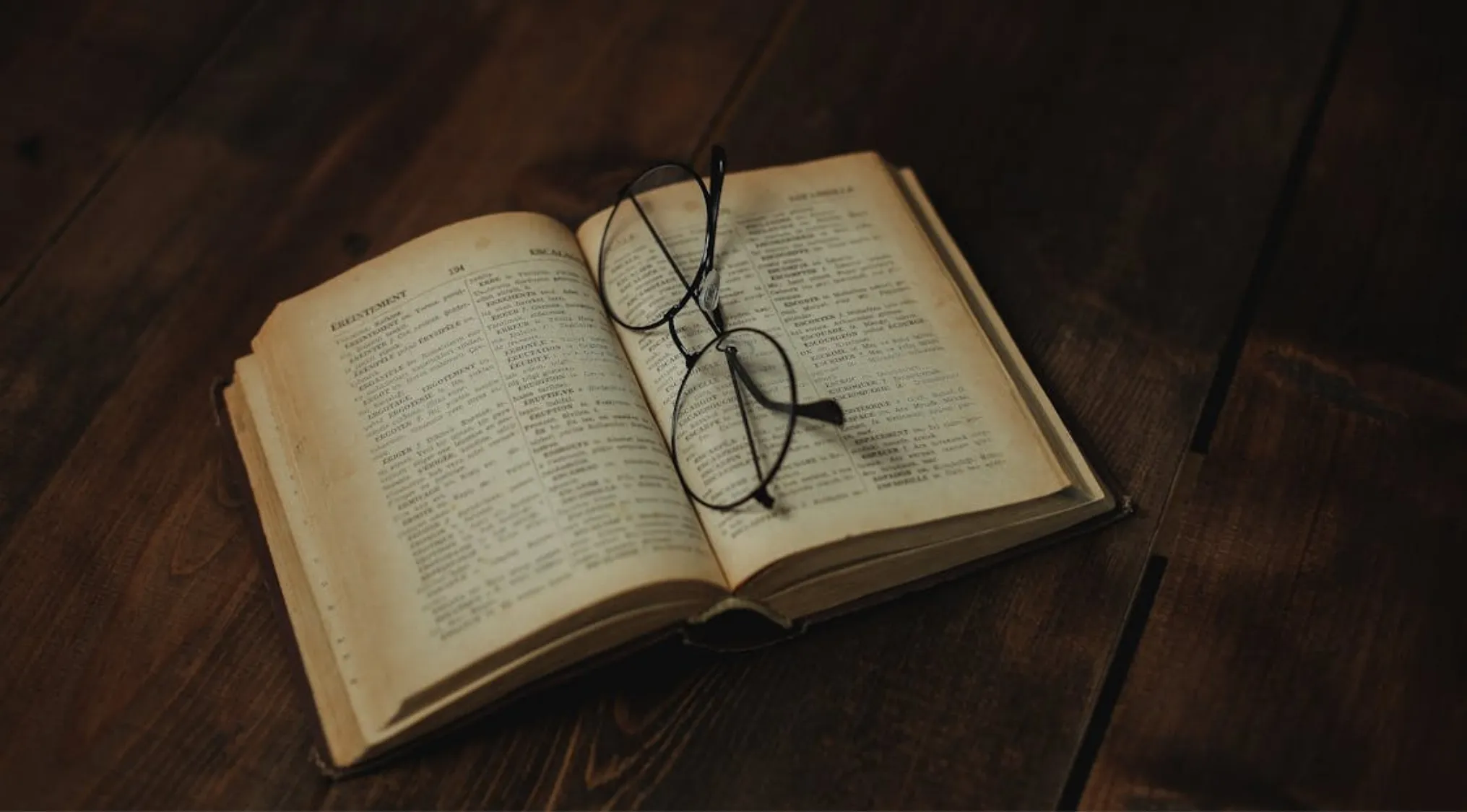 Open dictionary pages with reading glasses resting on top on a wooden surface, illustrating opposite meaning words.
