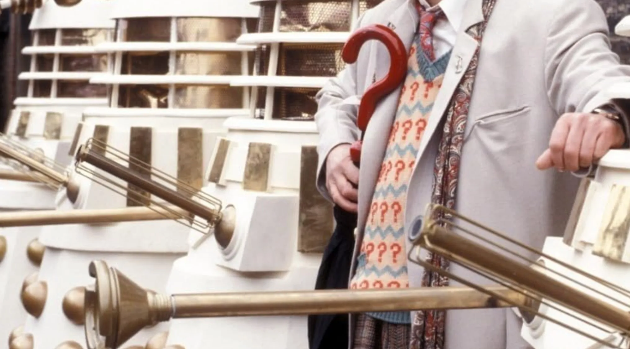 Man in vintage outfit holding a red question mark umbrella, standing next to white and gold sci-fi props from TV show.
