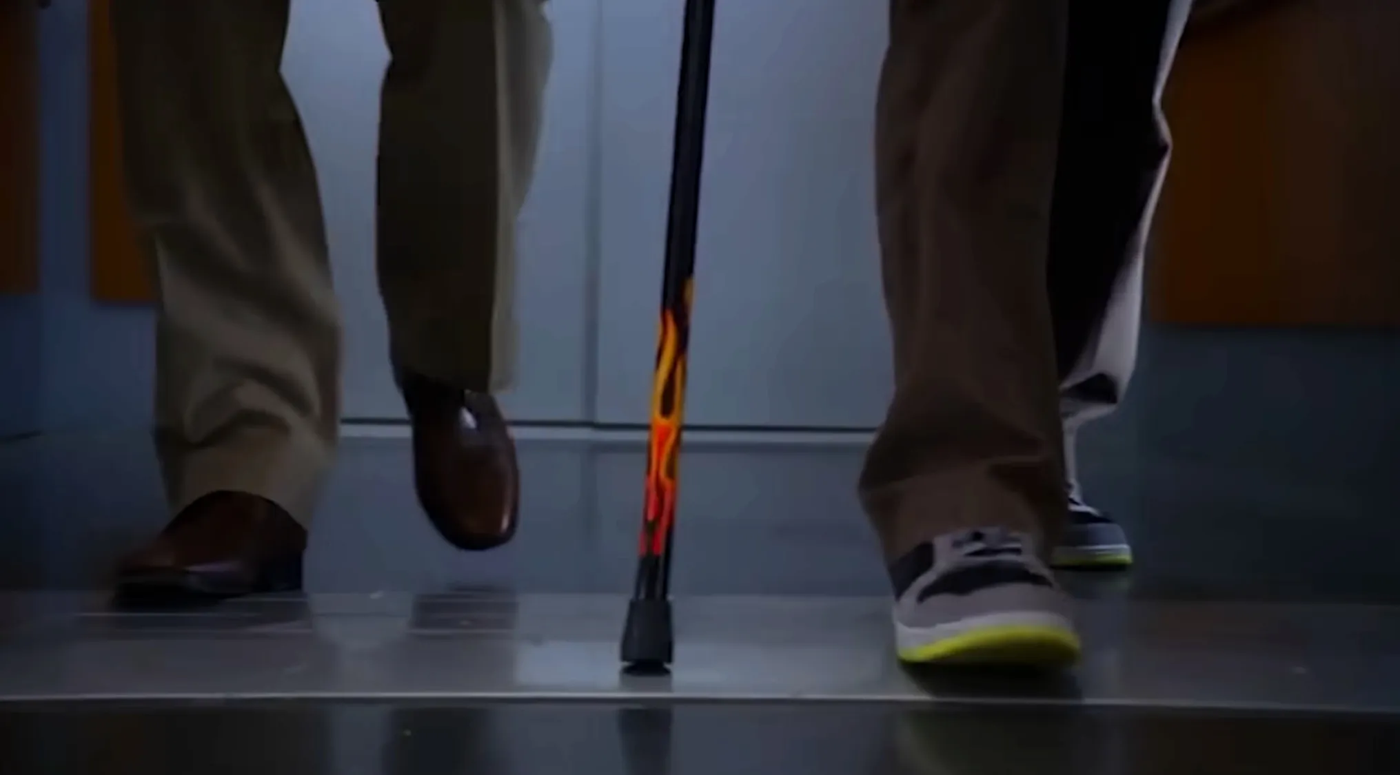 Close-up of a cane with flame design held by a TV show main character walking on a shiny floor with another person nearby.