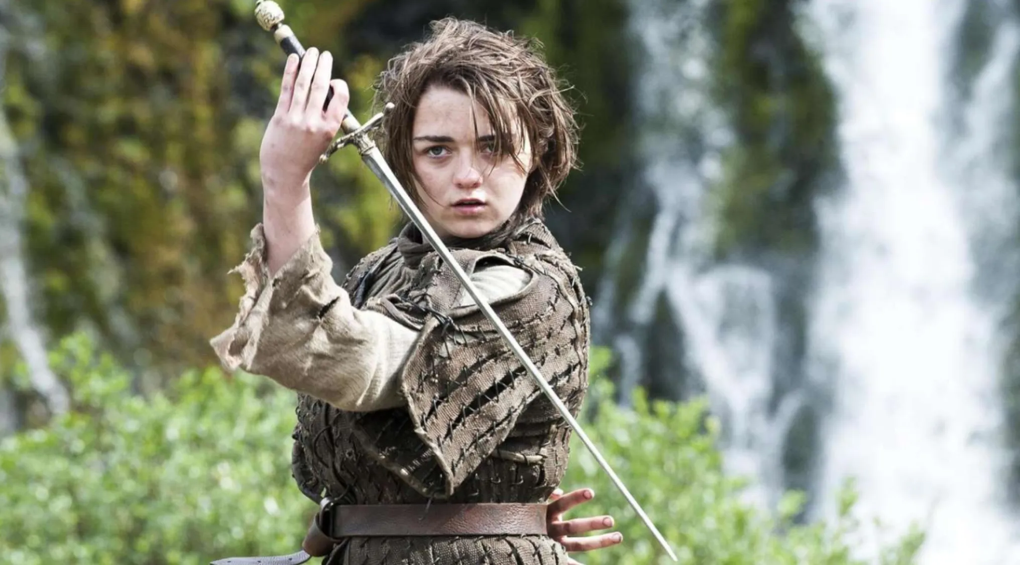 Character from Game of Thrones holding the iconic weapon, a sword, in a forested area with a waterfall background.