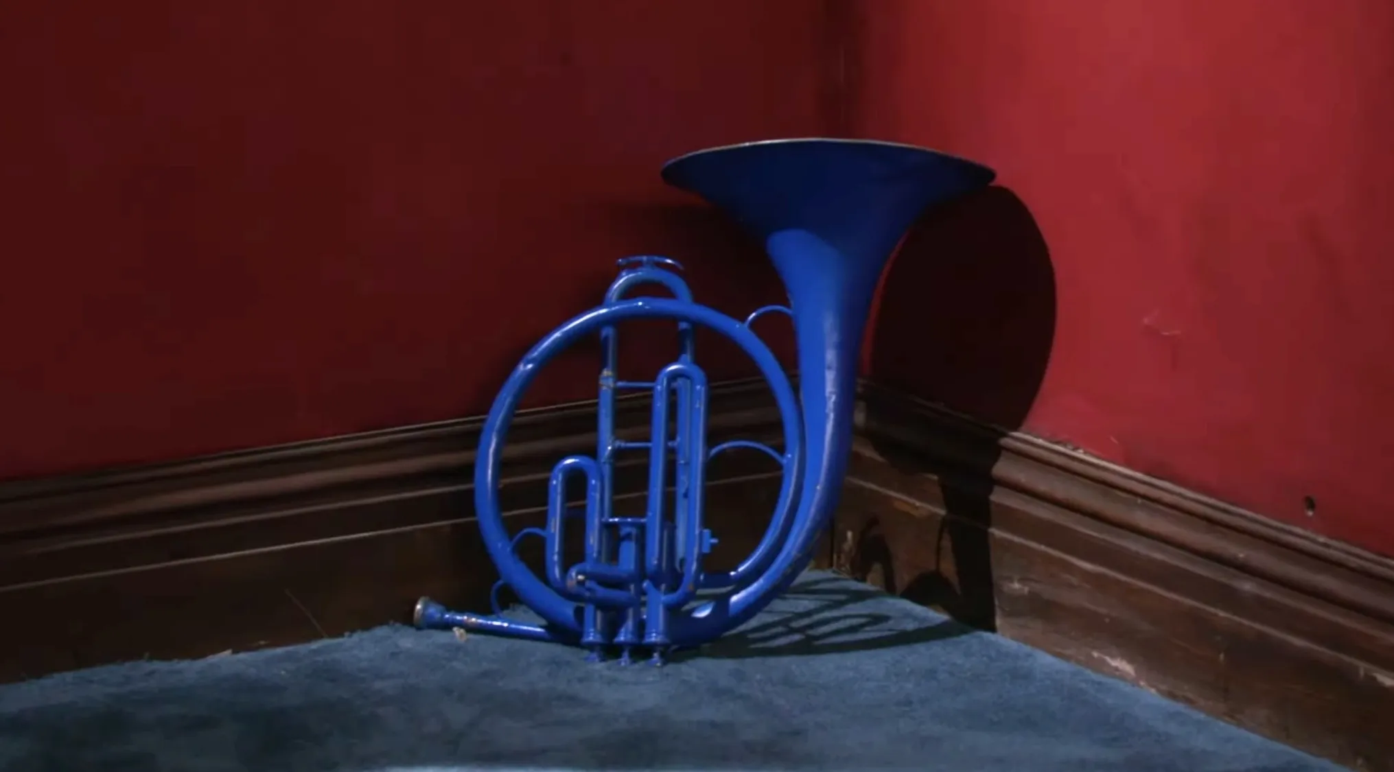 Blue brass musical instrument resting in a red corner of a room with carpet and wood trim in a TV show props quiz.