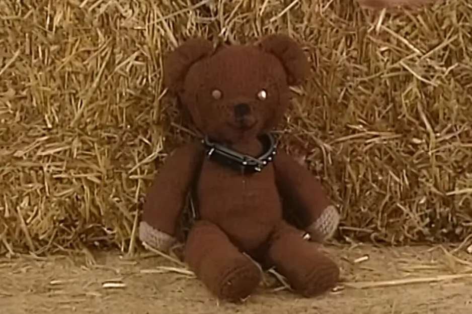 Brown teddy bear prop sitting against a haystack on a dirt surface with a collar around its neck.