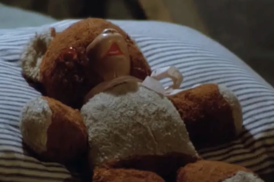 Teddy bear prop lying on a striped pillow in a dimly lit scene.