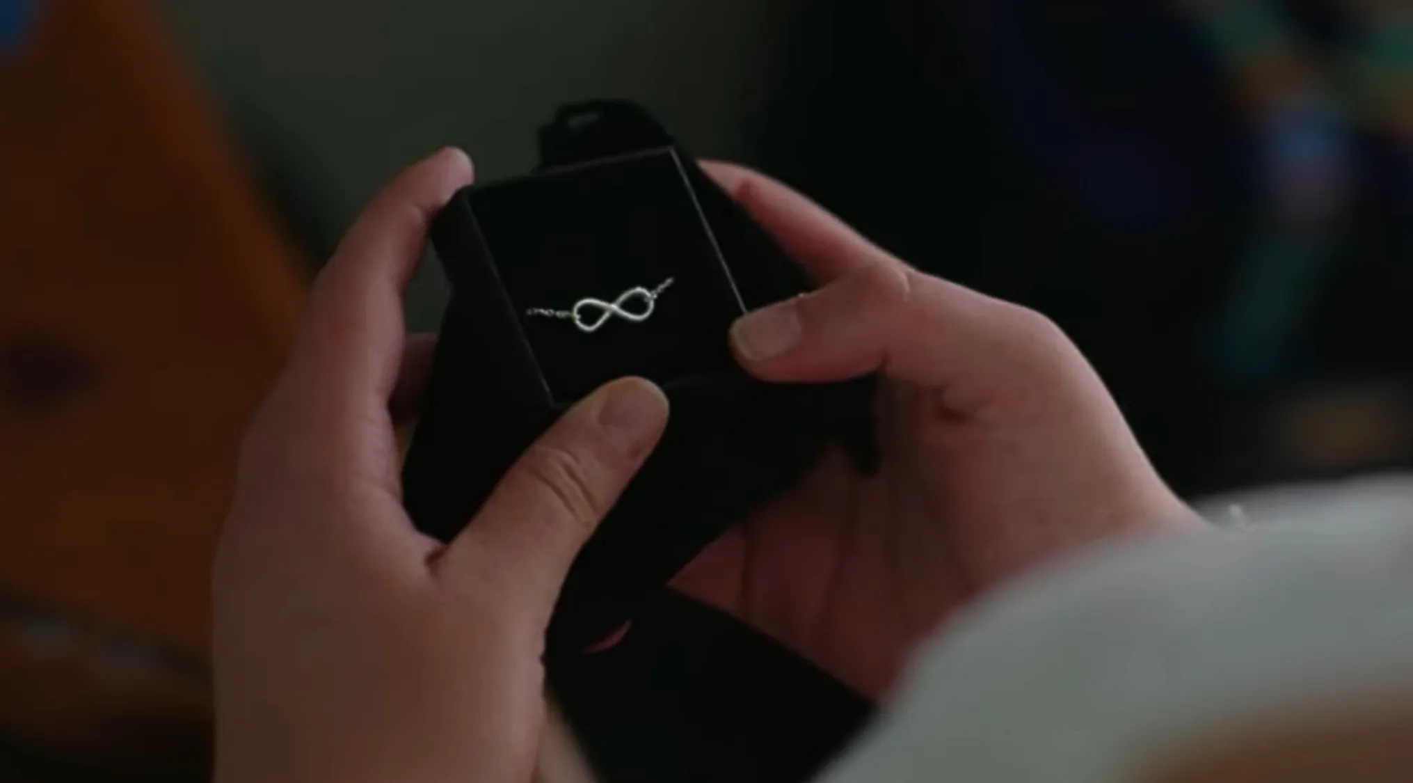 Hands holding a black box with a necklace featuring an infinity symbol in a TV show props quiz setting.