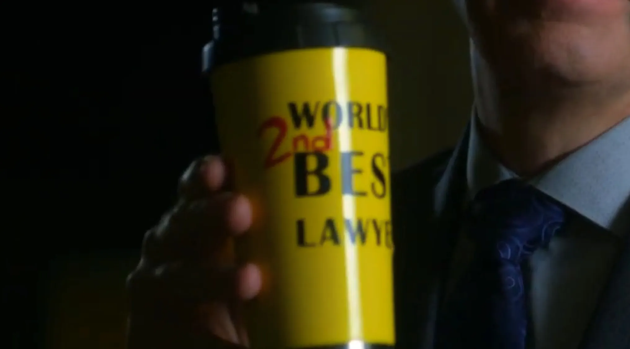 Man in suit holding a yellow travel mug with text about 2nd best lawyer in a TV show props quiz scene.