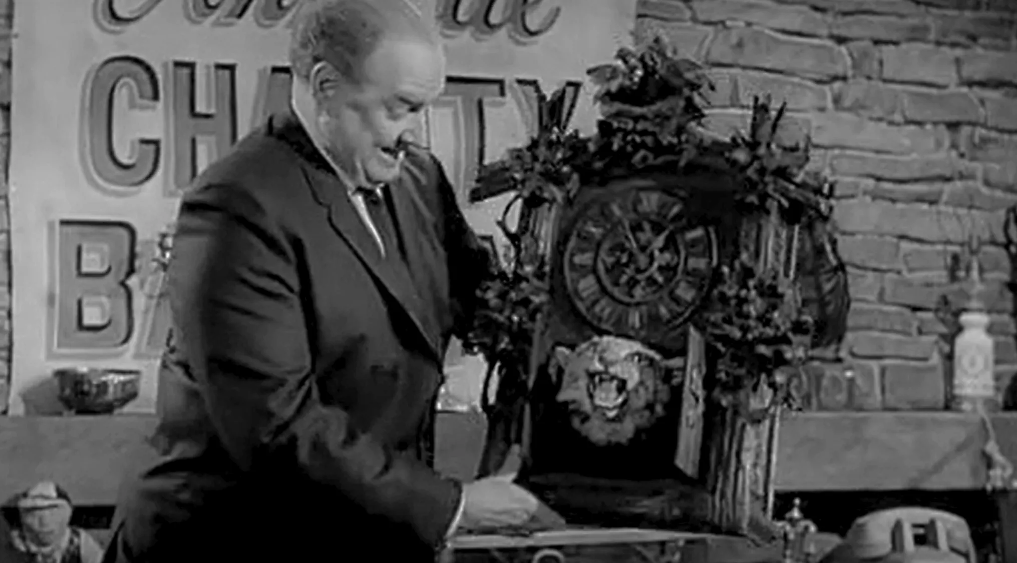 Black and white TV show scene featuring a man with a bizarre clock prop in a vintage studio setting.