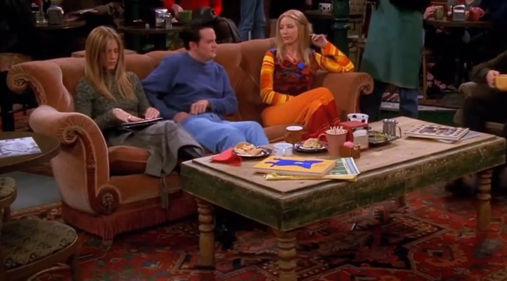 Friends TV show iconic coffee shop couch with characters sitting around a rustic wooden table in Central Perk cafe.
