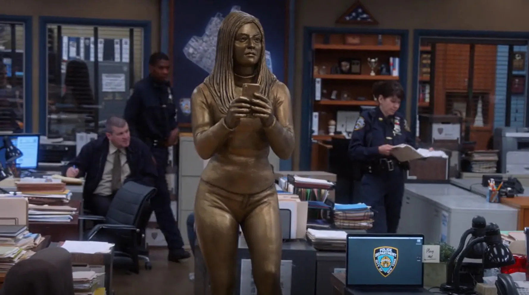 Bronze statue of a Brooklyn Nine-Nine character holding a phone in the precinct office surrounded by desks and officers.
