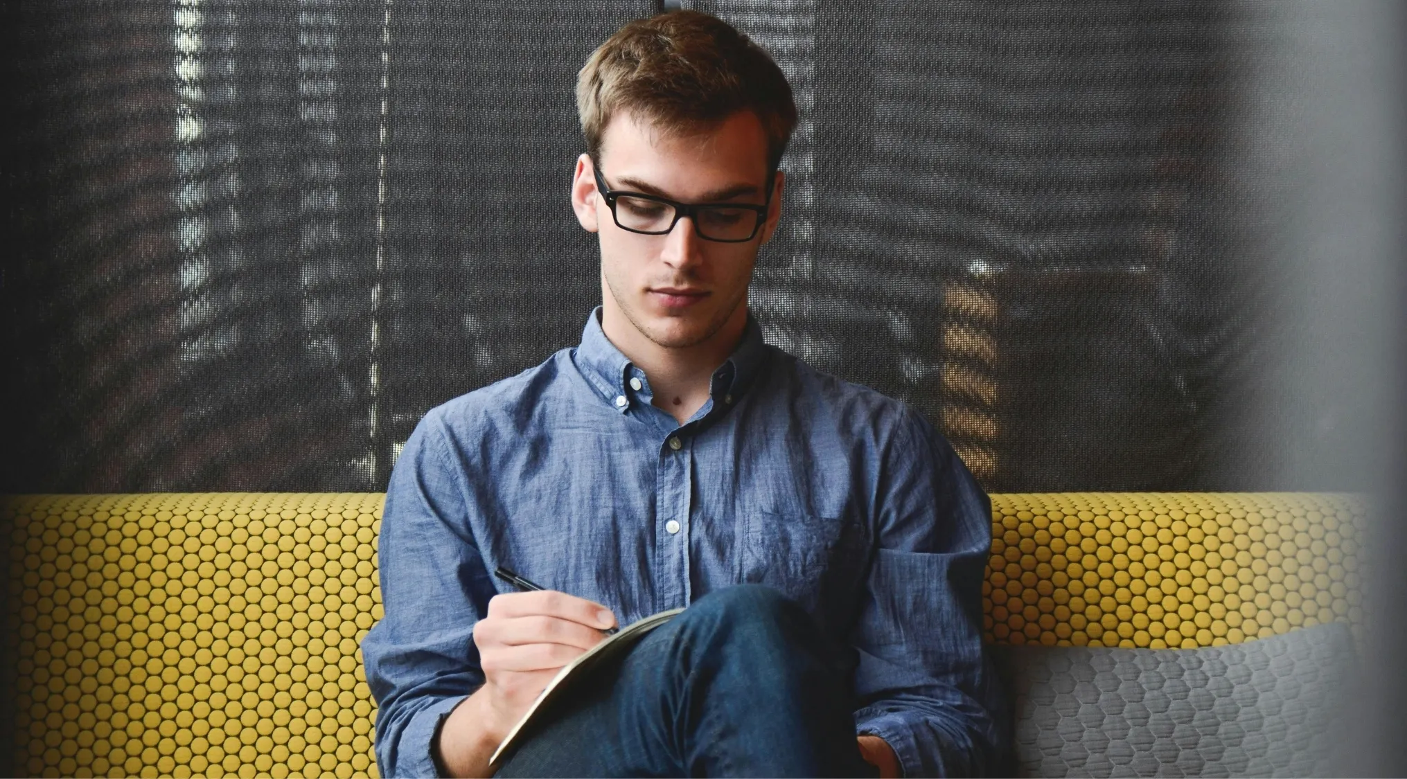 Young man wearing glasses, sitting on a patterned couch, focused on writing for a language quiz and standard English test.