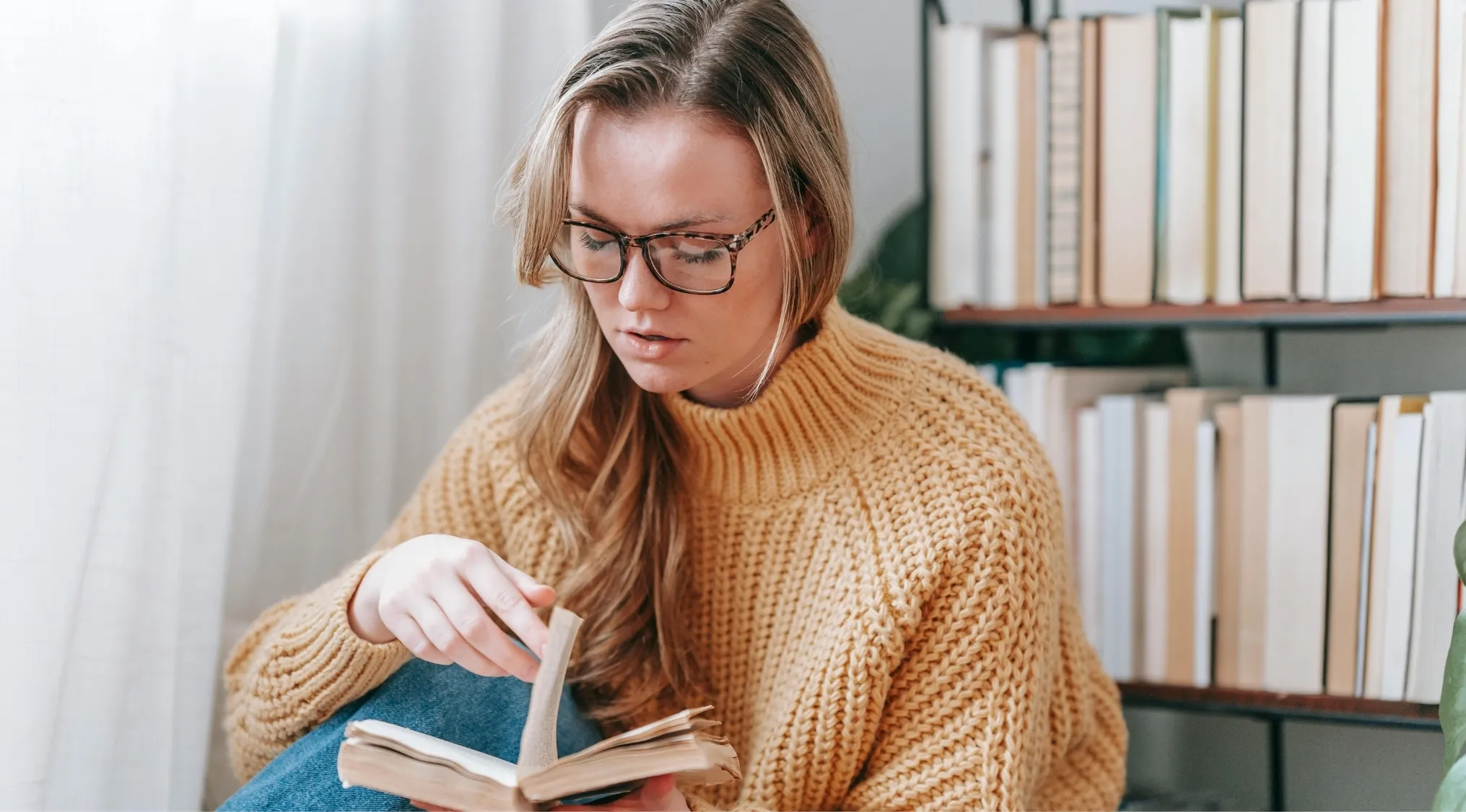 Young woman in glasses and a yellow sweater reading a book, testing her literary language skills in a cozy setting.