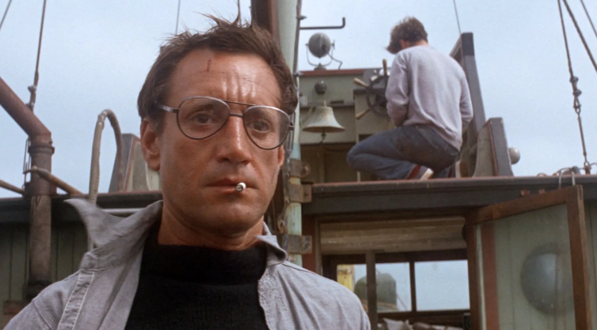 Close-up of a man smoking on a boat with another person in the background in a 70s movie scene.