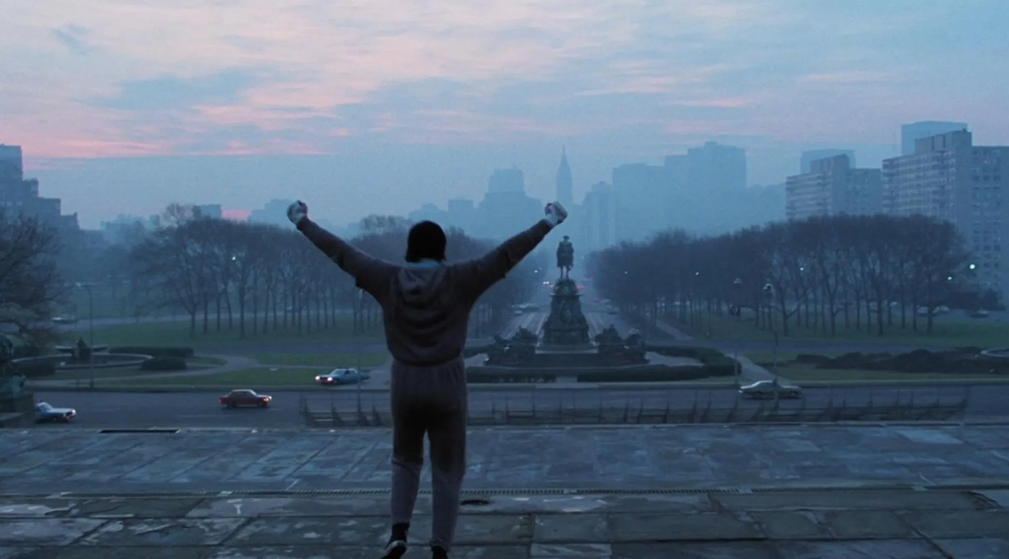 Man in tracksuit raising arms victoriously on steps overlooking city park and statue in a 70s movie scene.